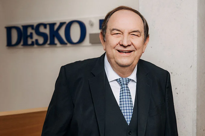 Un hombre mayor vestido de traje sonr&iacute;e en el pasillo de una oficina moderna, al fondo se ve el logotipo de DESKO.