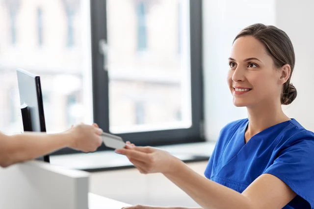 Mujer vestida de m&eacute;dico acepta una tarjeta de una persona en un mostrador de recepci&oacute;n.