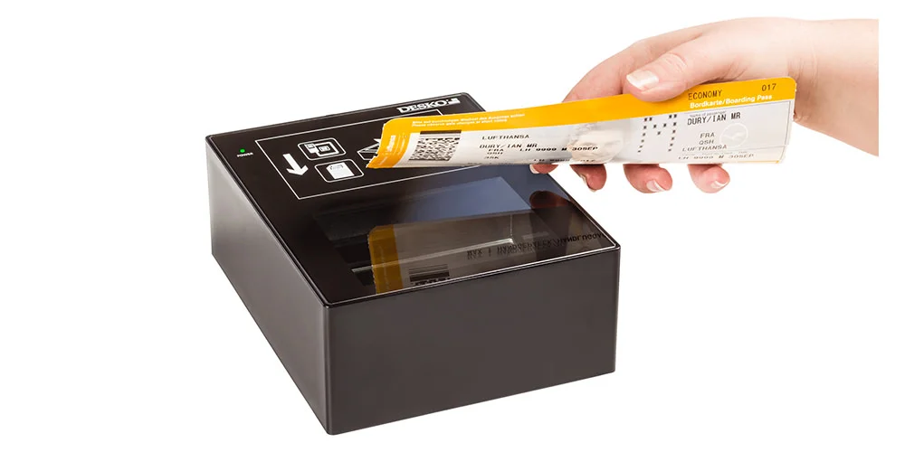 Mano sosteniendo tarjeta de embarque amarilla sobre un esc&aacute;ner de documentos DESKO compacto con superficie de cristal.