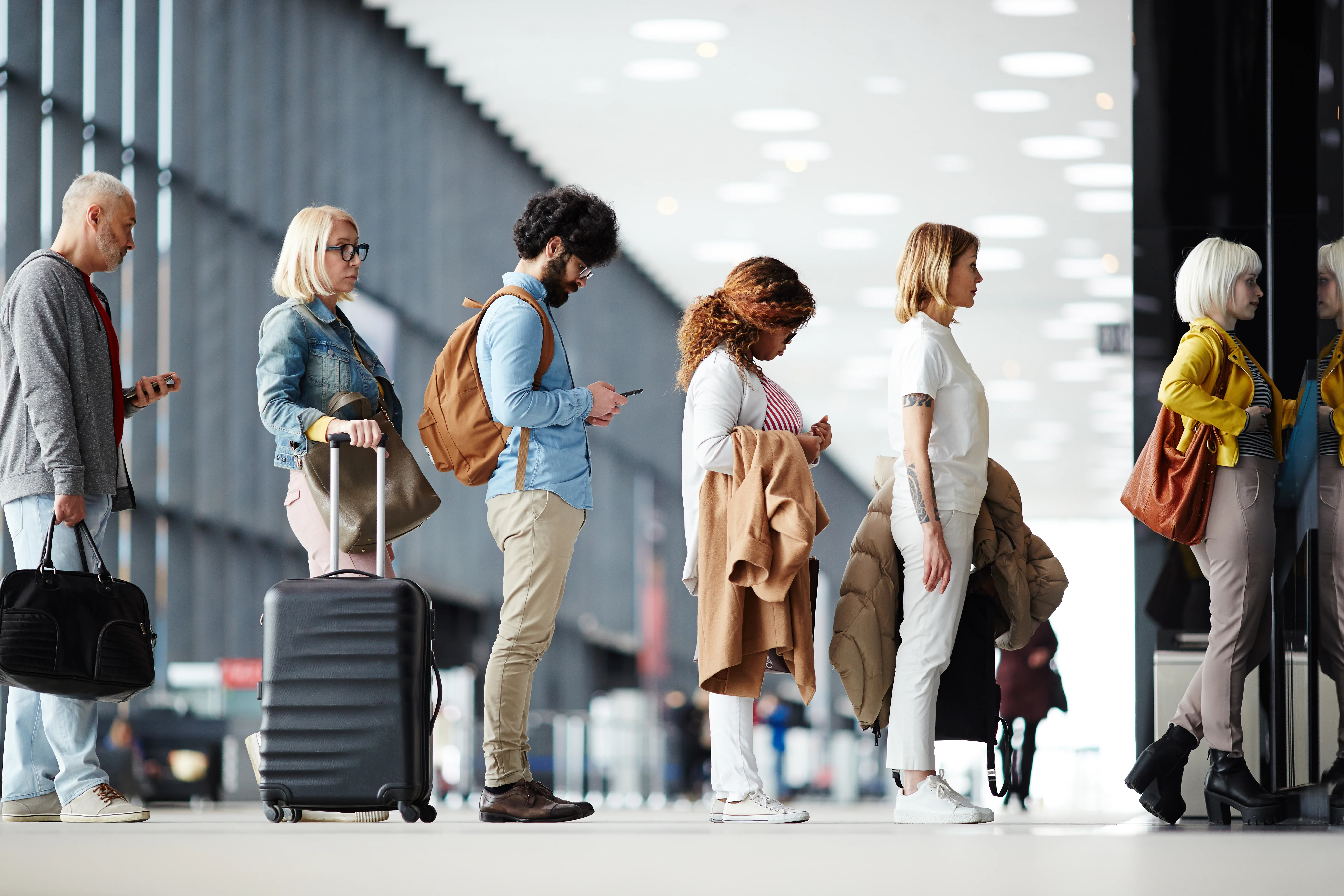 Varios viajeros hacen cola en la moderna terminal del aeropuerto, algunos con maletas y los ojos puestos en sus smartphones.