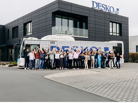 Gran grupo de empleados animando delante del moderno edificio de la empresa DESKO y el autocar con la marca en las instalaciones de la empresa.
