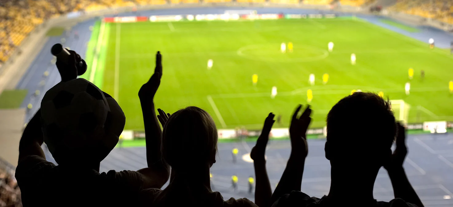 Siluetas de espectadores animando con los brazos levantados miran desde las gradas un partido de f&uacute;tbol en un estadio iluminado.