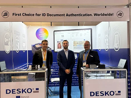 在 DESKO 展览会的展台上，三个人站在文件扫描仪和显示器前，上面写着 