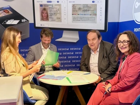 Cuatro personas en el stand de la feria DESKO durante una situación de consulta con una comprobación de documentos y una demostración de software al fondo.