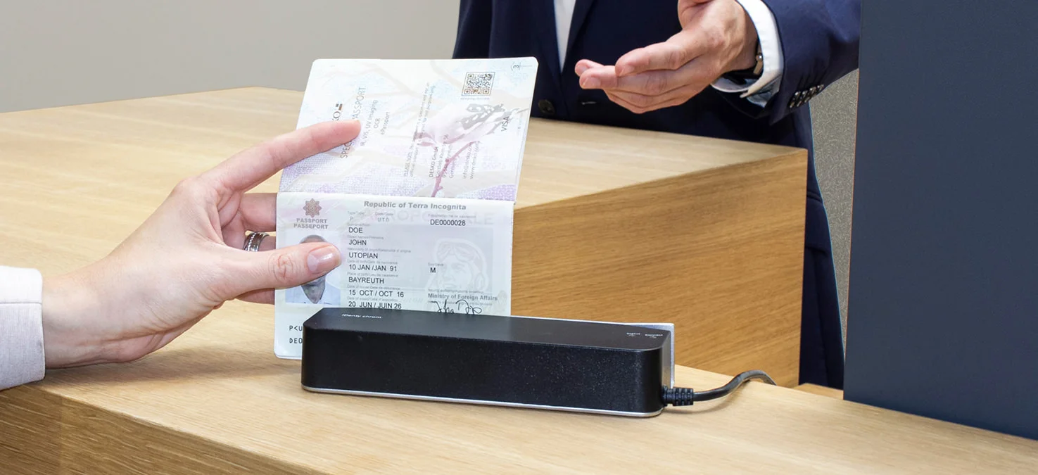 Una mano sostiene un pasaporte abierto sobre un lector en el mostrador mientras un empleado detr&aacute;s le pide que compruebe su identidad.