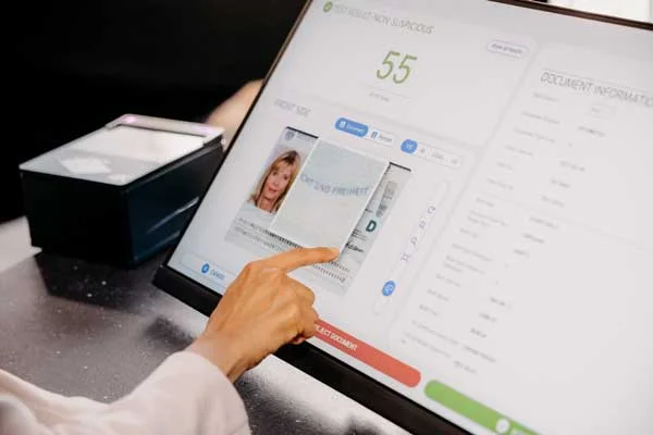La persona se&ntilde;ala un an&aacute;lisis de identificaci&oacute;n digital en una pantalla, al lado hay un lector de documentos en el mostrador.