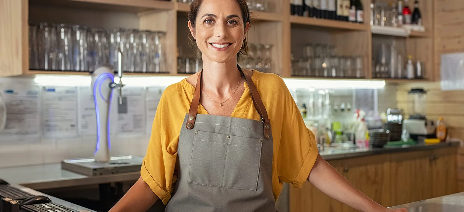 Personal de servicio amable con delantal de pie detr&aacute;s de un mostrador en una cafeter&iacute;a o bistr&oacute;, con vasos y botellas de bebidas al fondo.