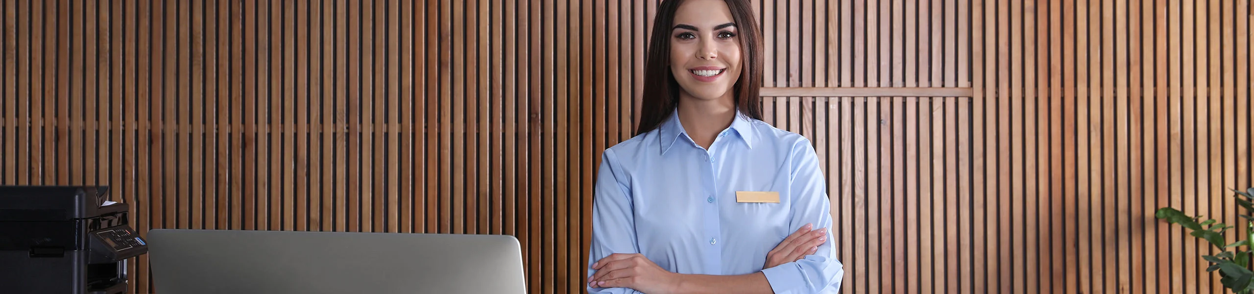 Una amable recepcionista con una placa identificativa est&aacute; de brazos cruzados detr&aacute;s de un mostrador frente a una pared de lamas de madera.
