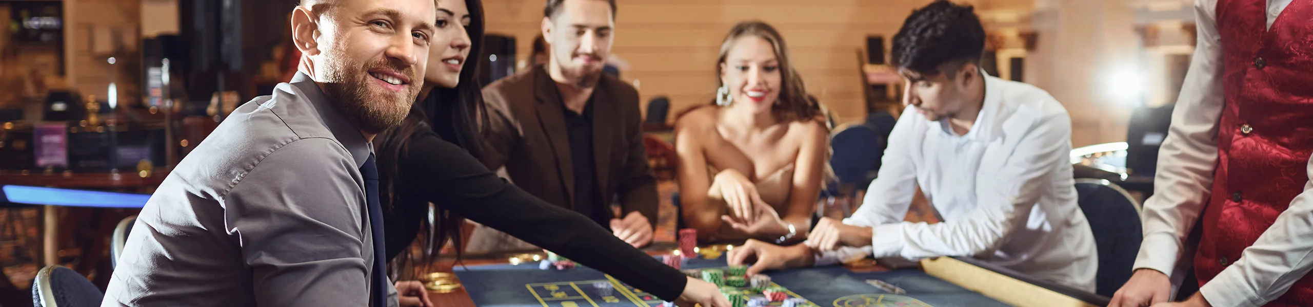 Grupo de personas elegantemente vestidas sentadas a una mesa de juego en un casino, colocando fichas y cartas y jugando juntas en un ambiente cordial.