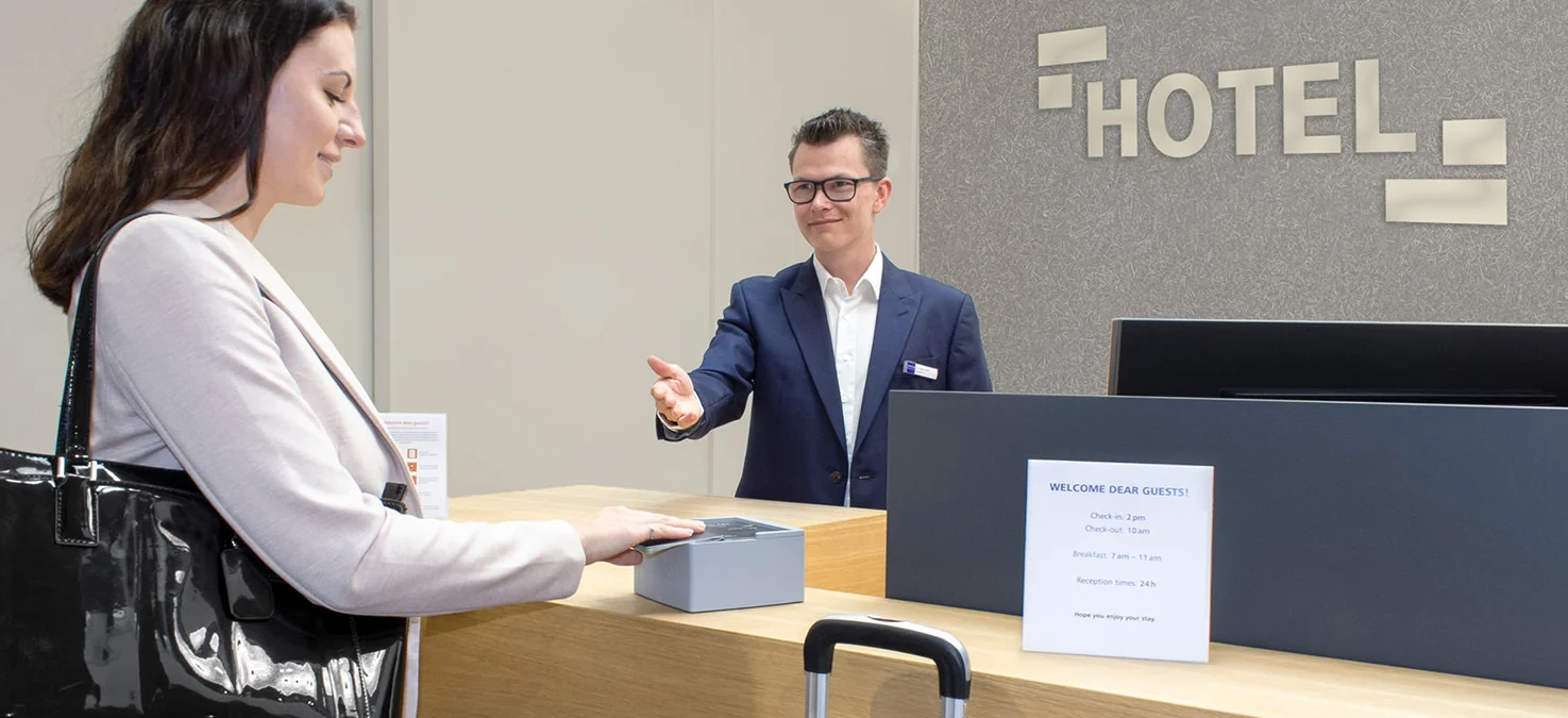 El hu&eacute;sped del hotel coloca una tarjeta en un lector sin contacto en el mostrador de recepci&oacute;n mientras un empleado le ayuda detr&aacute;s del mostrador.