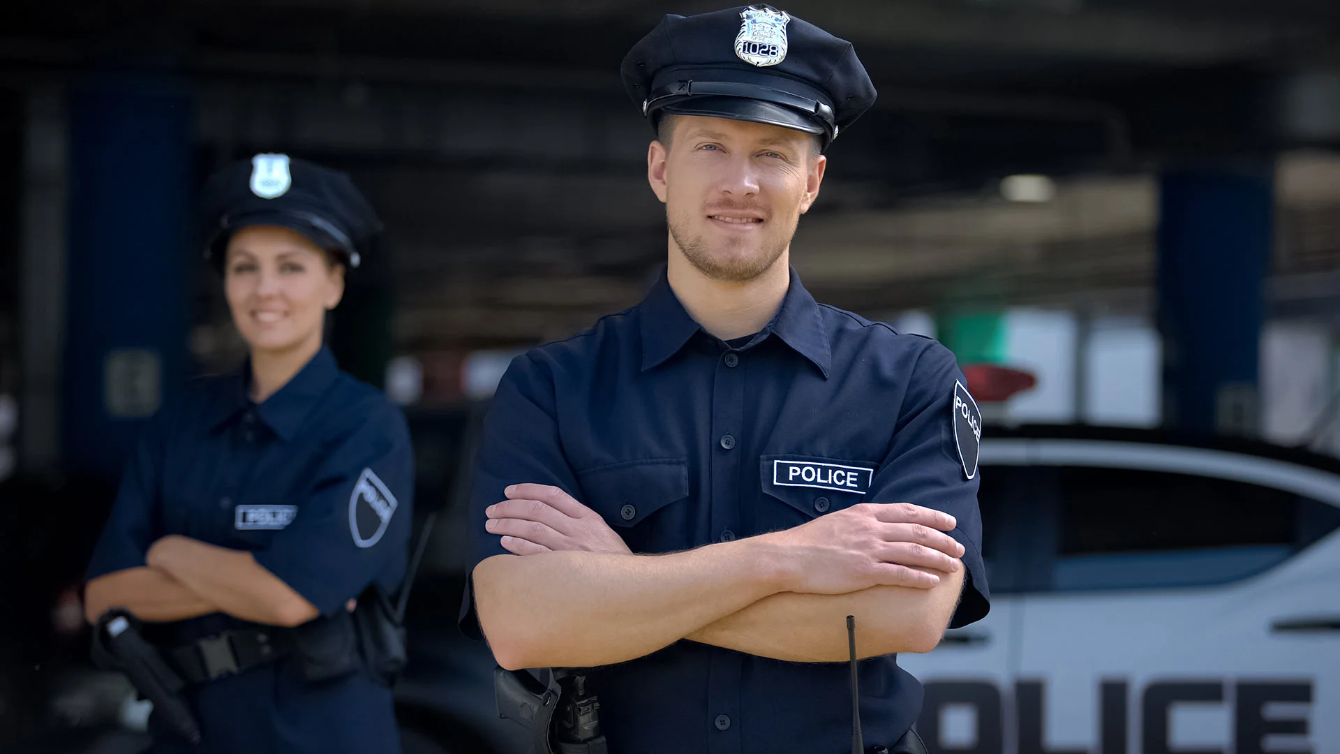 Ein Polizist und eine Polizistin stehen mit verschr&auml;nkten Armen vor einem Polizeiauto.