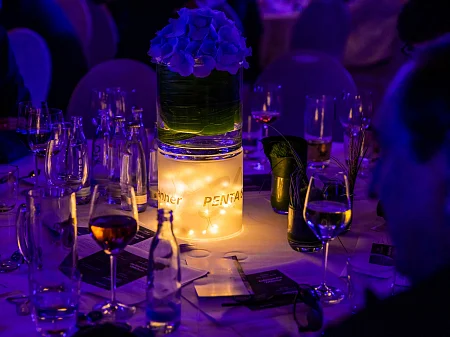 Mesa festiva con cilindro de cristal iluminado, arreglo floral, velas y la marca PENTA en el evento nocturno.