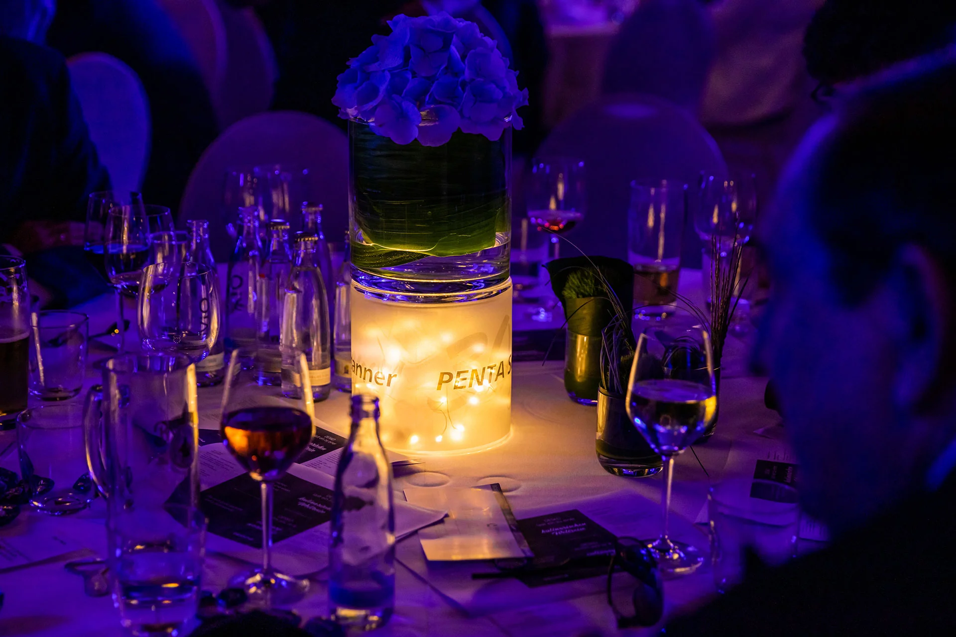 Mesa festiva con cilindro de cristal iluminado, arreglo floral, velas y la marca PENTA en el evento nocturno.