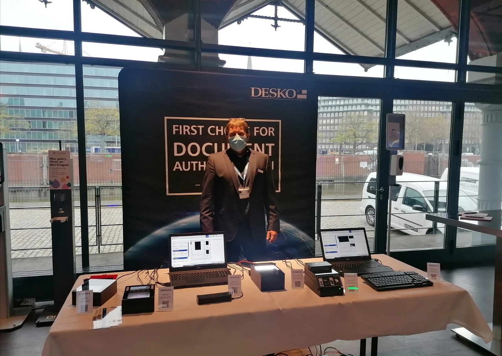 Stand de feria con la marca DESKO, empleado con m&aacute;scara presenta esc&aacute;ner, ordenadores port&aacute;tiles y tecnolog&iacute;a de pruebas sobre mesa.