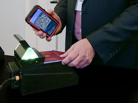 La mano sujeta un smartphone con un código QR, mientras el pasaporte reposa sobre un escáner de identificación iluminado para su inspección.