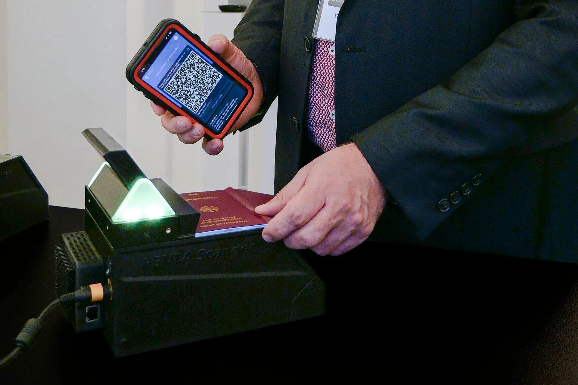 La mano sujeta un smartphone con un c&oacute;digo QR, mientras el pasaporte reposa sobre un esc&aacute;ner de identificaci&oacute;n iluminado para su inspecci&oacute;n.