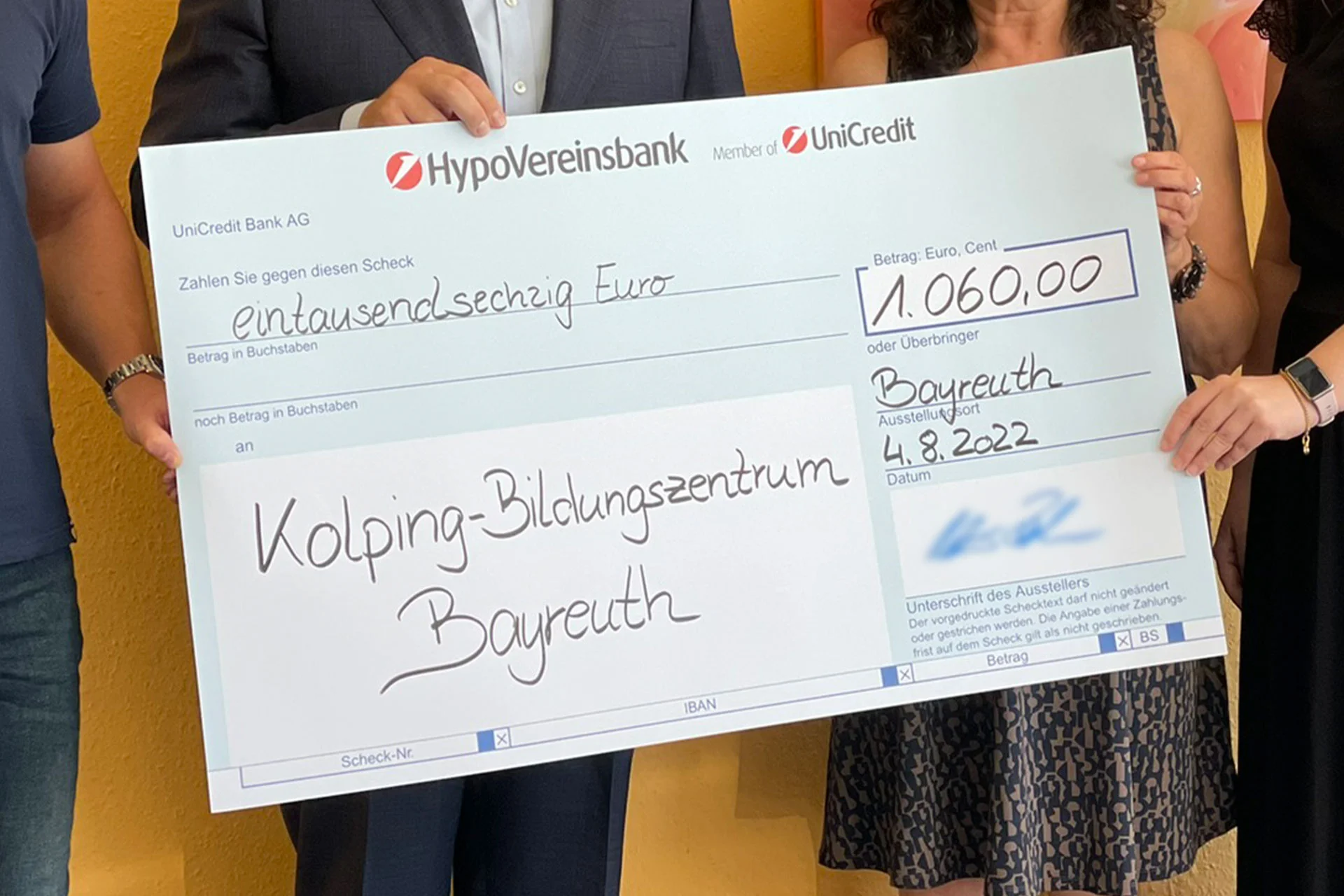 Varias personas sostienen un gran cheque donativo de 1.060 euros para el Centro de Educaci&oacute;n Kolping de Bayreuth.