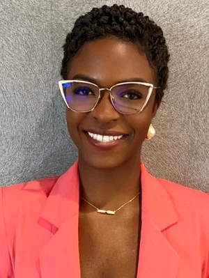 Retrato de mujer sonriente con peinado corto, gafas claras y americana rosa sobre fondo neutro.